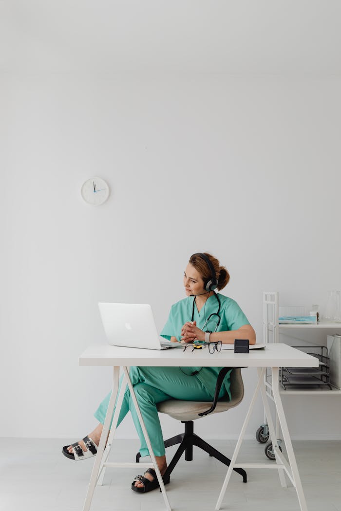 Healthcare professional in scrubs using laptop for telemedicine session in modern office.